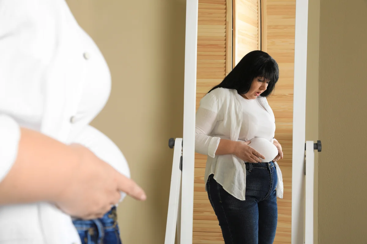 A woman standing in front of a mirror, observing her belly, highlighting the need for exercises to lose belly fat at home.