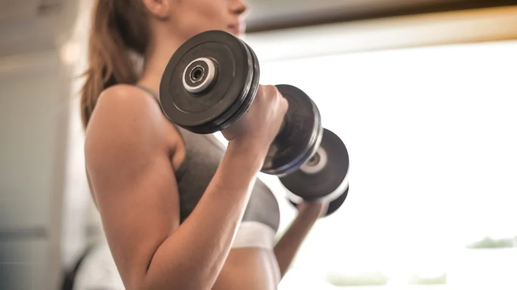 A focused woman lifting dumbbells during a strength workout, showcasing her toned arms and commitment to fitness in a bright, modern indoor setting.