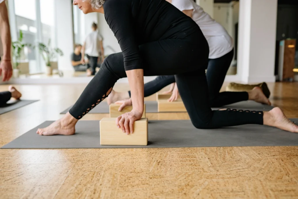 Participants perform gentle stretches in a lunge position using wooden blocks for support during a yoga or rehabilitation session, emphasizing balance and flexibility.