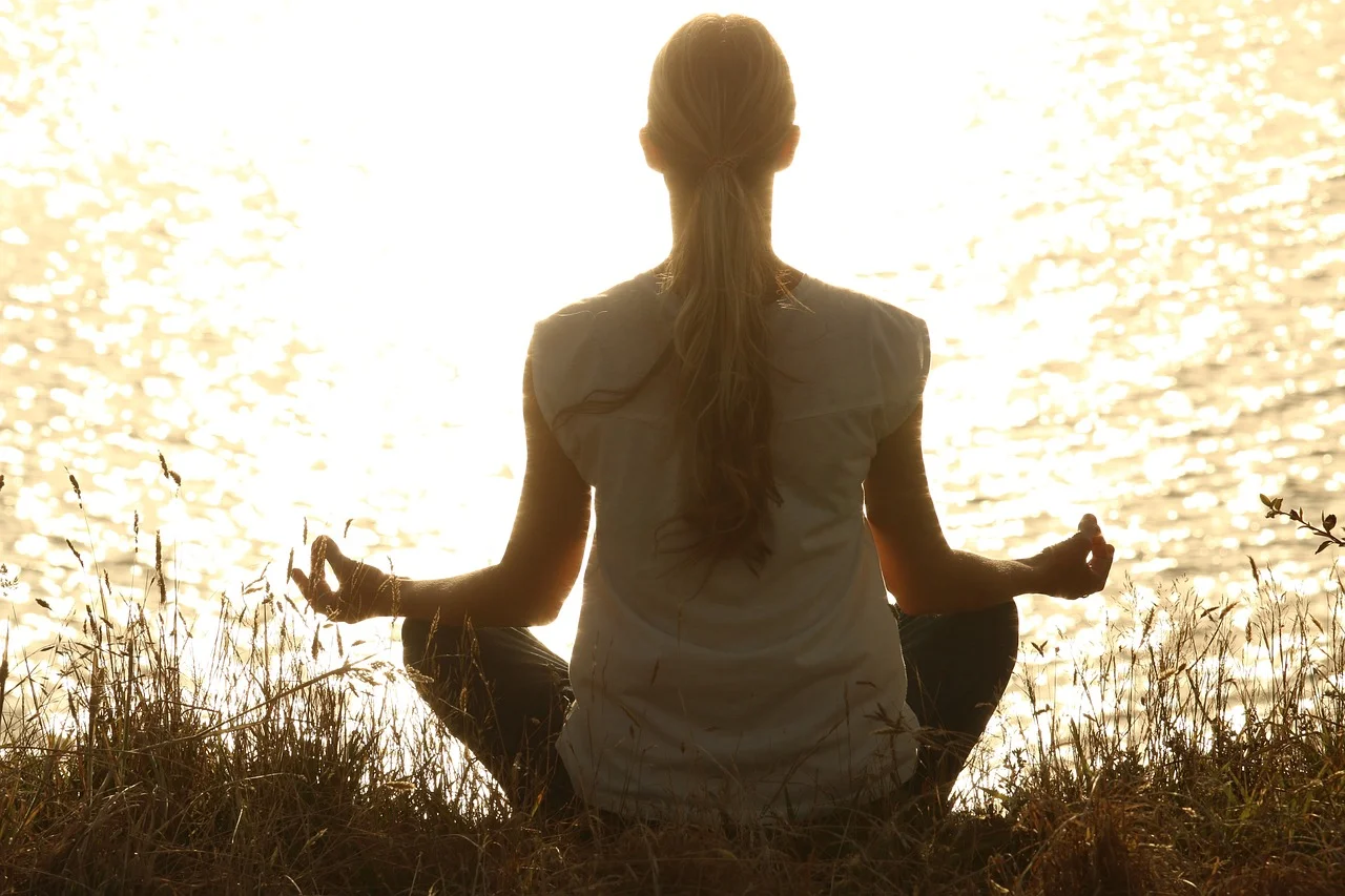 Woman meditating outdoors by a lake, symbolizing the connection between meditation and exercise to enhance focus, recovery, and overall workout effectiveness.