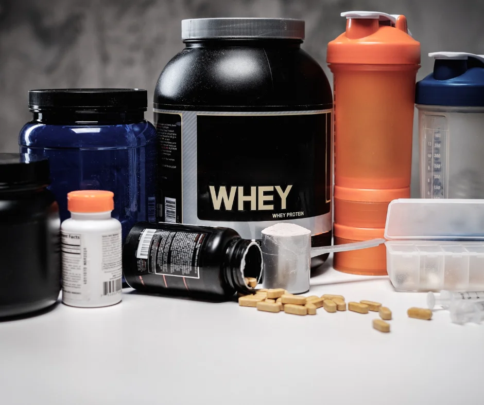 A collection of fitness supplements including a large container of whey protein, vitamin bottles, and shaker bottles, alongside scattered supplement pills and scoops.