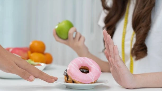 A person holding an apple while rejecting a plate with a pink-frosted donut, symbolizing the choice of healthy foods over sugary snacks.