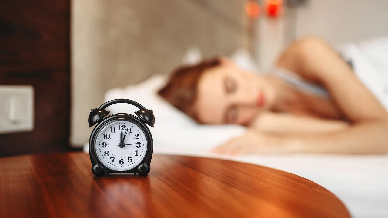 Is Going to Bed Hungry Helps to Lose Weight? A woman peacefully sleeping in bed with an alarm clock showing 12:00 placed on a bedside table. The image suggests a restful night, relevant to discussions about the effects of going to bed hungry on sleep quality and weight loss.