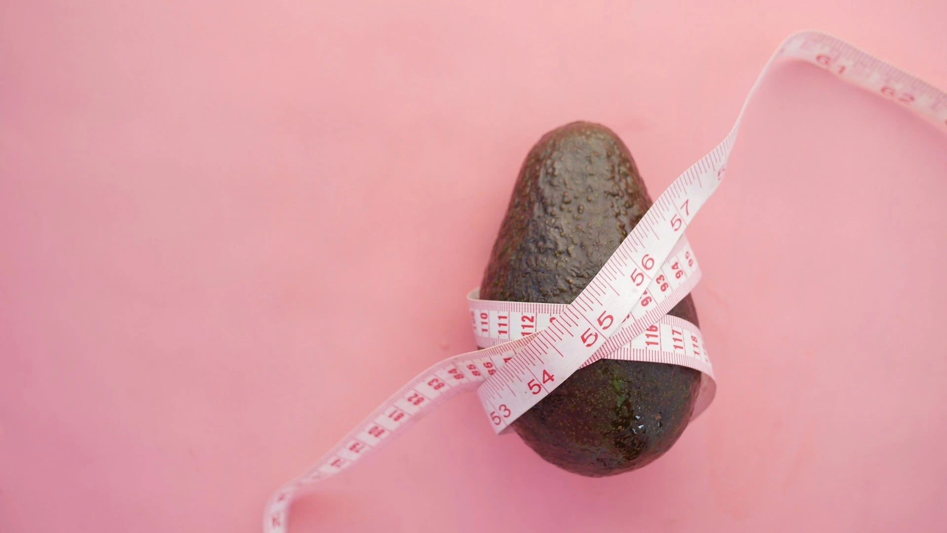 An avocado wrapped with a measuring tape on a pink background, symbolizing healthy eating and portion control in 'the menopause diet 5 day plan to lose weight.'