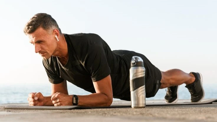 Man performing bodyweight forearm exercises outdoors on a mat, building grip strength and endurance. Wearing black sportswear with a water bottle nearby.