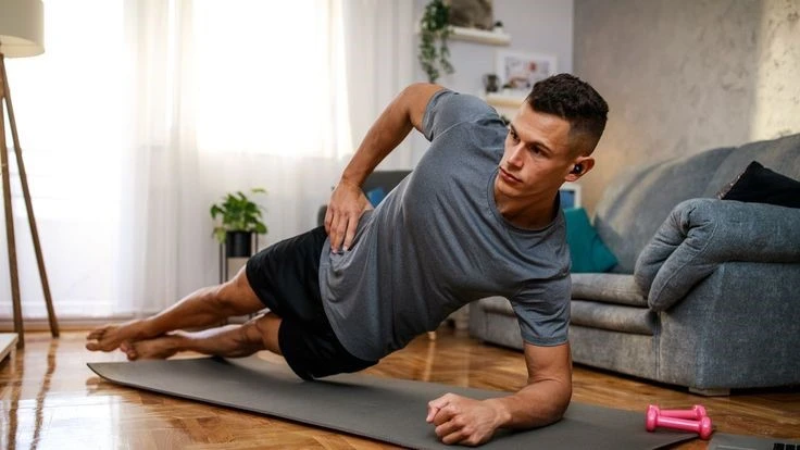 A fit man performing a side plank as part of his bodyweight pull exercises routine at home. He is wearing a gray t-shirt and black shorts, balancing on a yoga mat in a cozy living room setting with a couch and dumbbells nearby.