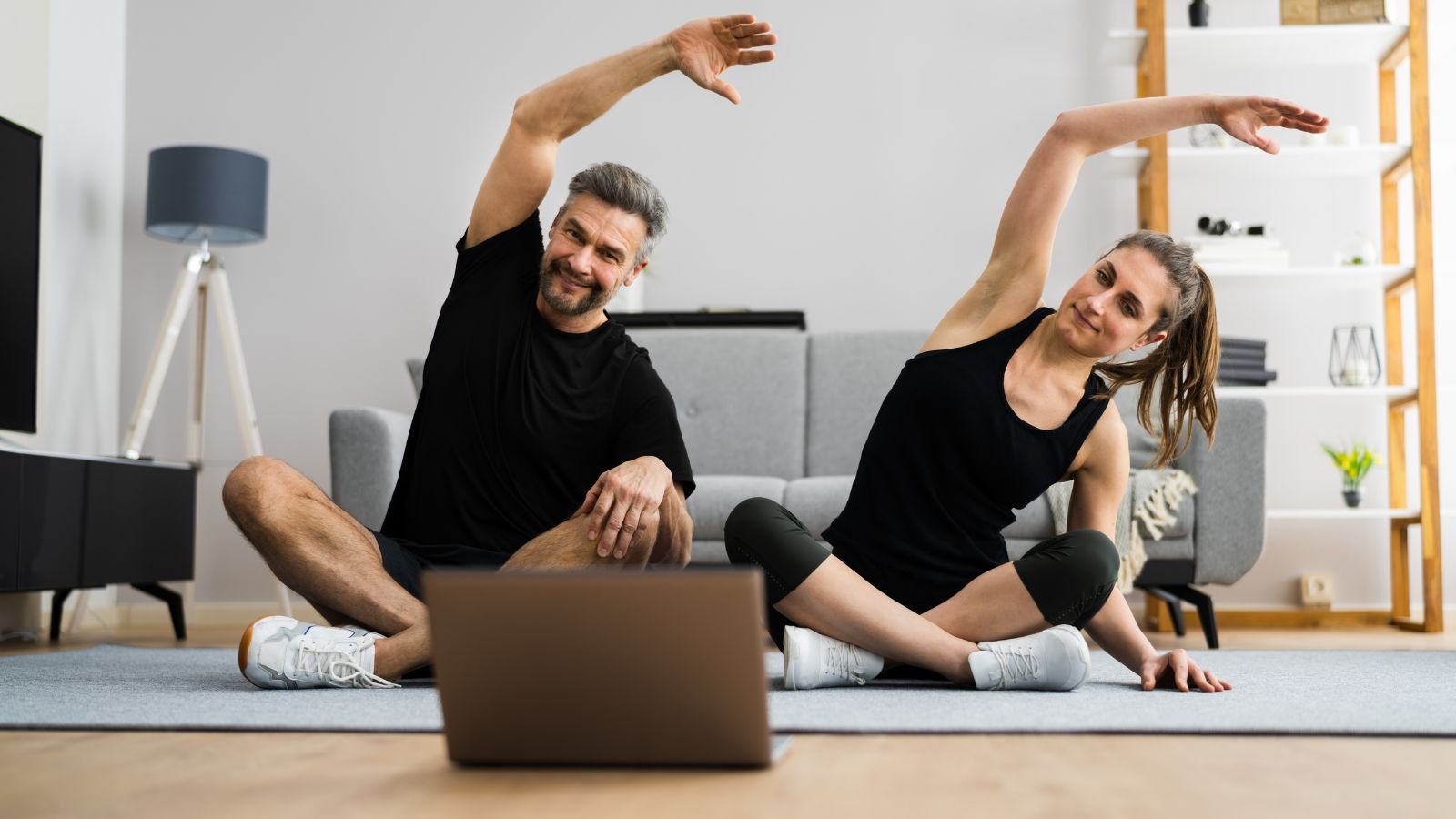 Workouts for Busy Professionals – A man and woman in athletic wear doing a home workout, stretching together in front of a laptop, making fitness easy and accessible from home.