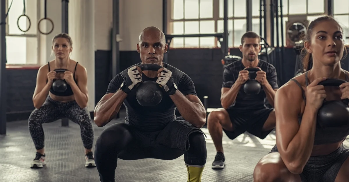 A group of men and women performing Goblet Squats with kettlebells in a gym, demonstrating proper squat form and posture for strength training.
